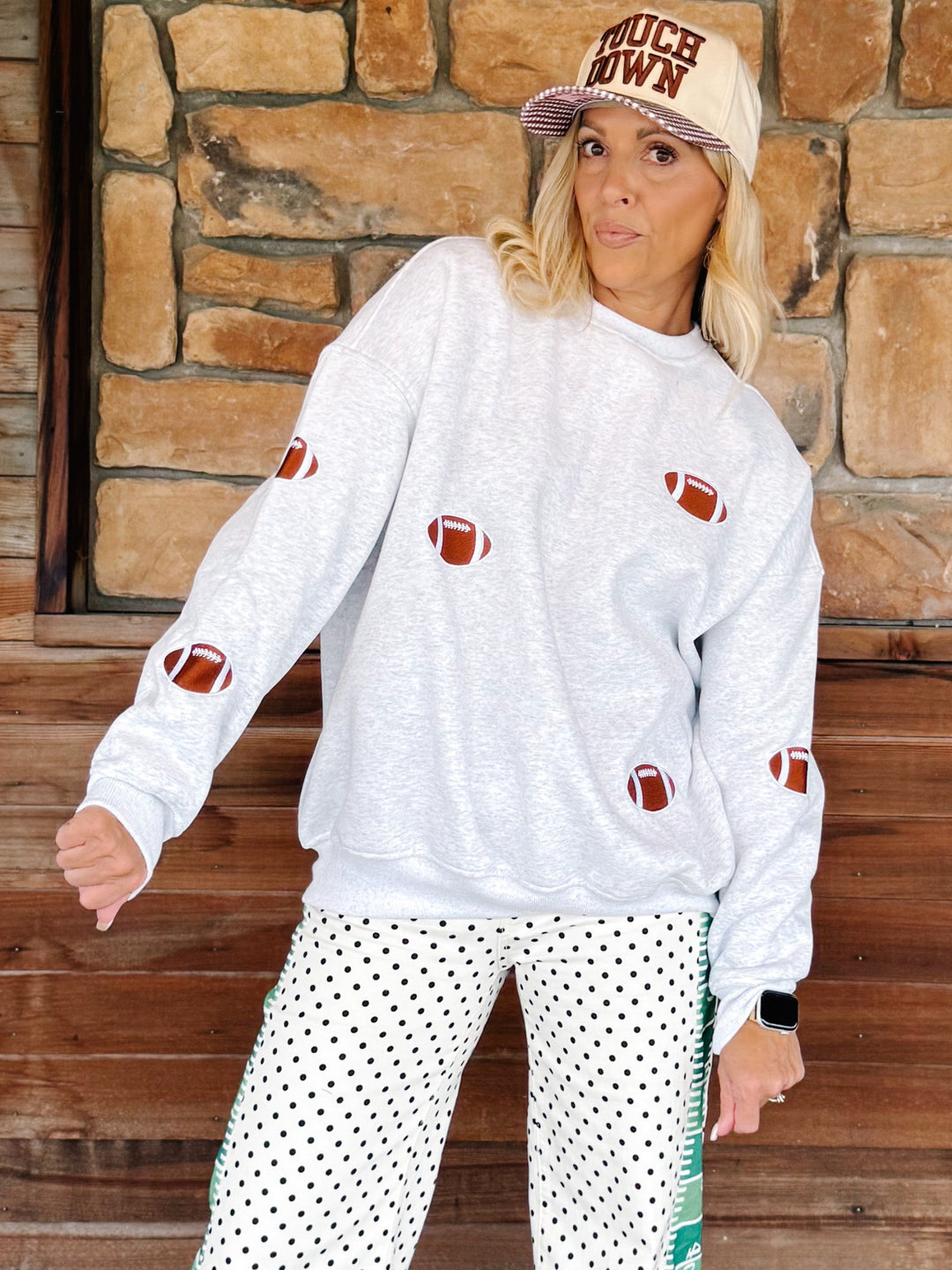 A woman modeling a white crewneck sweatshirt with brown football graphics, paired with white pants featuring black polka dots and green side stripes, standing in front of a rustic stone wall.
