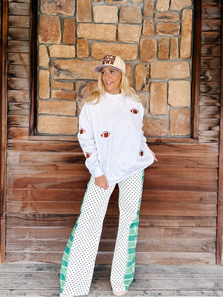 A woman stands in front of a rustic stone and wood wall wearing a white sweatshirt with football patch details, white wide-leg pants with green striped side panels and black polka dots, and a beige baseball cap that reads &