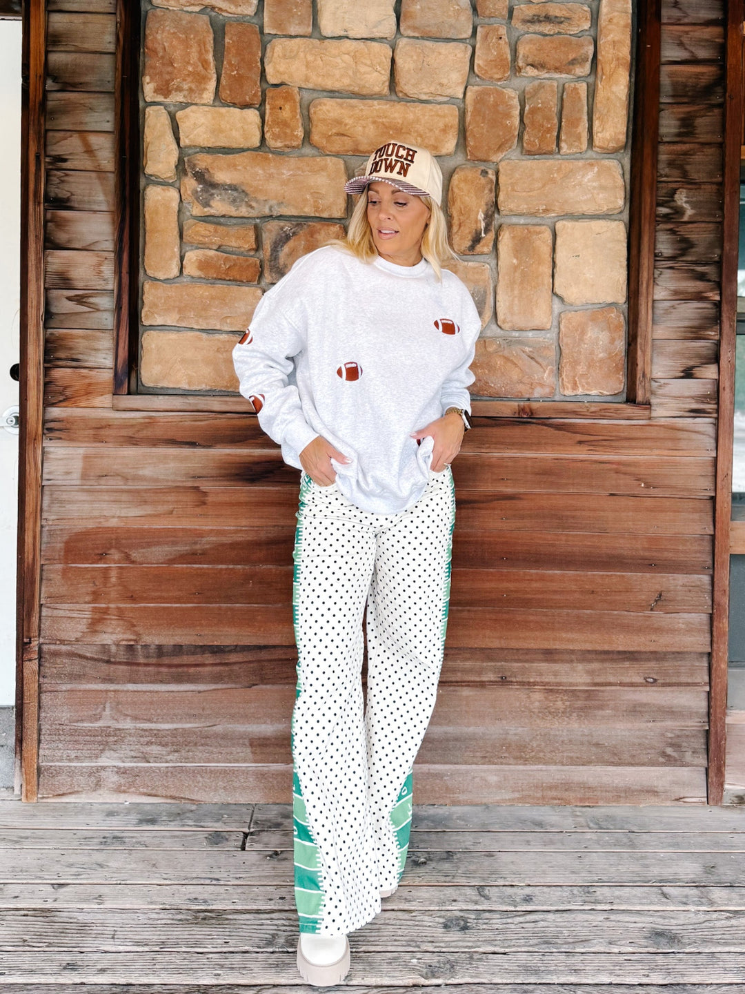 A woman stands on a rustic wooden porch in front of a stone wall, wearing a white sweatshirt with small brown football patches, white wide-leg pants with black polka dots and green side panels, and a beige cap.
