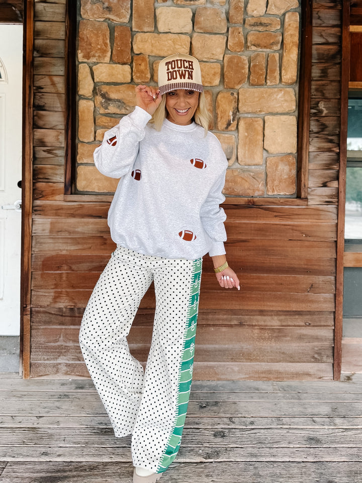A woman stands on a wooden porch in front of a stone wall, wearing a light gray long-sleeve sweatshirt with football motifs, white wide-leg pants with green side stripes and white polka dots, and a beige cap with &