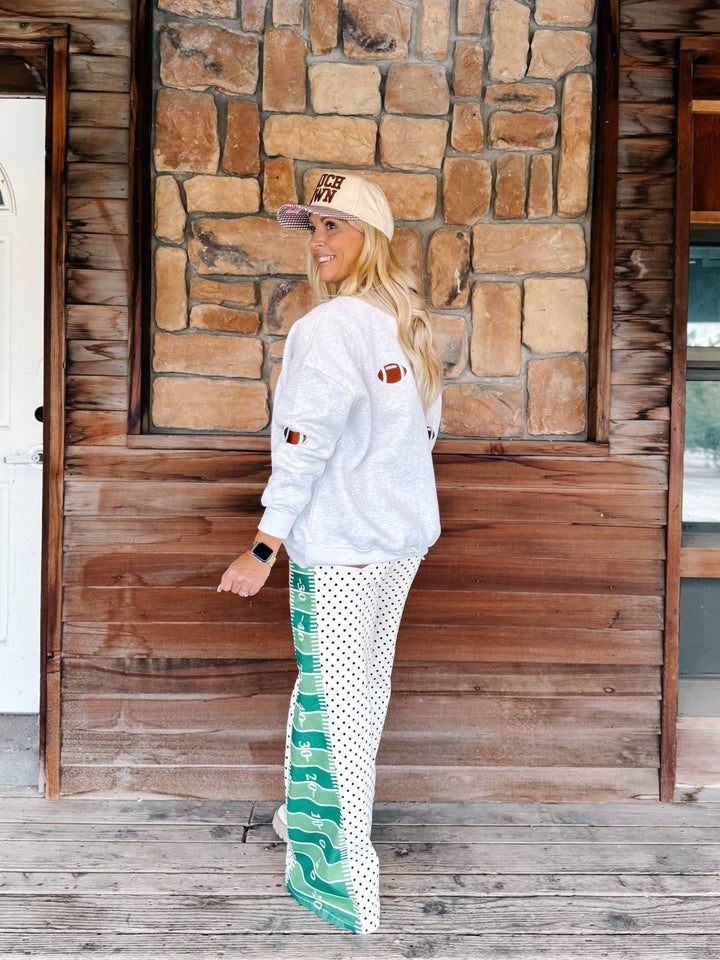 A woman stands on a rustic wooden porch in front of a stone wall, wearing a white sweatshirt with a football patch on the sleeve, a beige cap, and white pants with green and black polka dots and a green wave pattern near the hem.