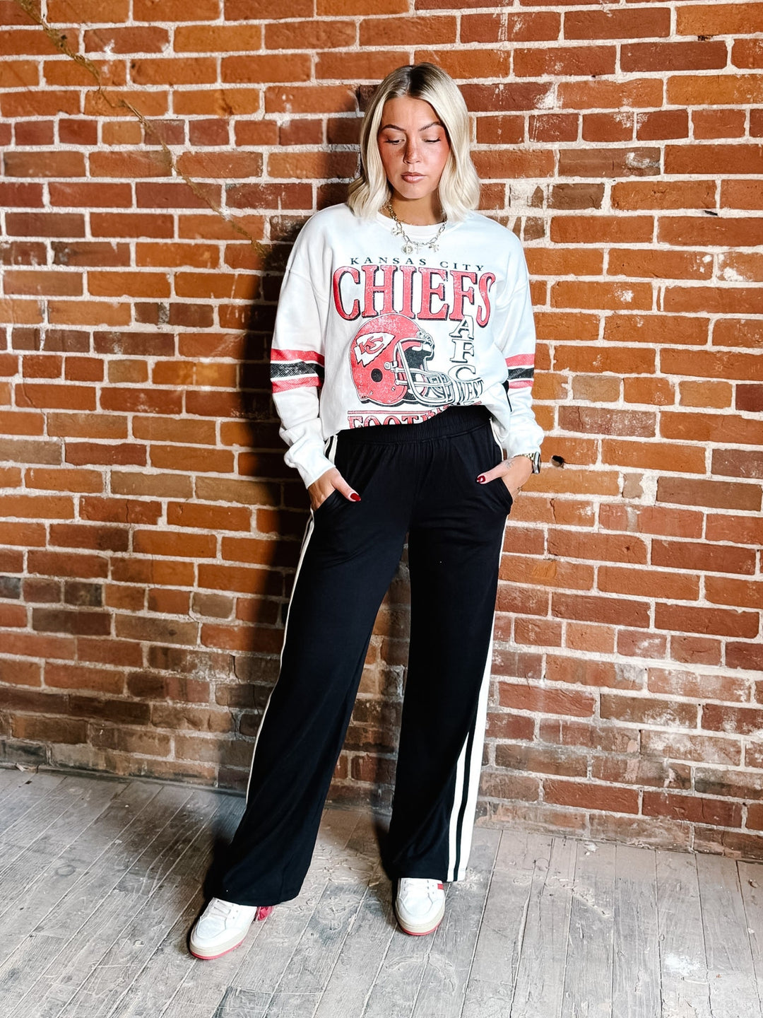 A woman stands against a red brick wall wearing a white Kansas City Chiefs sweatshirt with red and navy details, paired with black track pants featuring white side stripes and white sneakers.