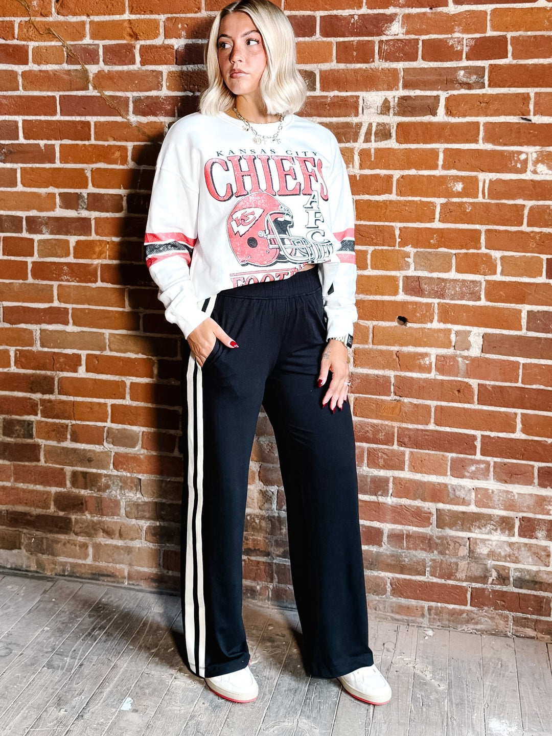 A woman stands against a red brick wall wearing a white graphic sweatshirt with red and pink &