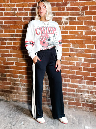 A woman stands against a red brick wall wearing a white graphic sweatshirt with red and pink 'CHIEFS' lettering and a football helmet print, paired with black wide-leg track pants featuring white side stripes, and white sneakers.