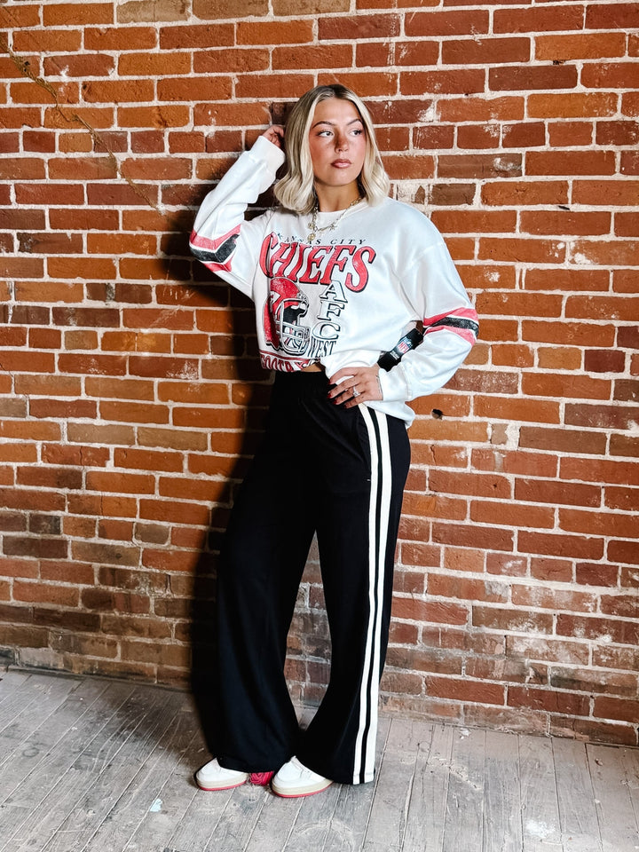 A woman posing in front of a brick wall wearing a white graphic sweatshirt with red and black details, black wide-leg track pants with white side stripes, and white sneakers.