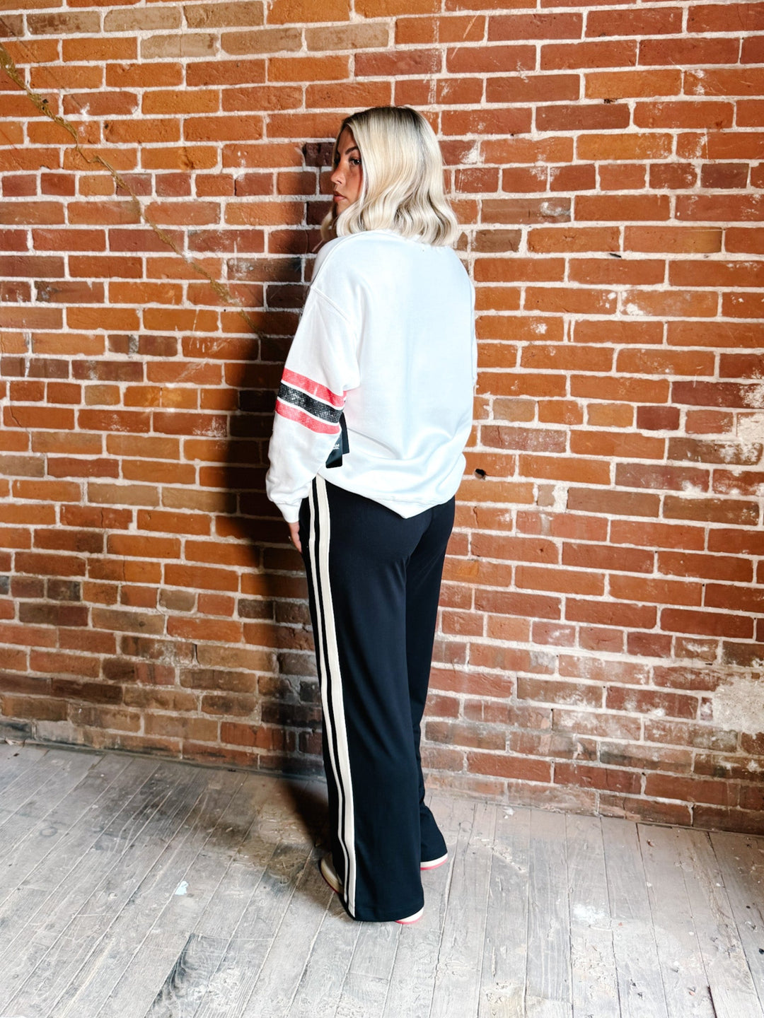 A woman stands with her back to the camera against a red-brown brick wall, wearing a white sweatshirt with pink and black stripes on the sleeves and black wide-leg track pants with white side stripes.