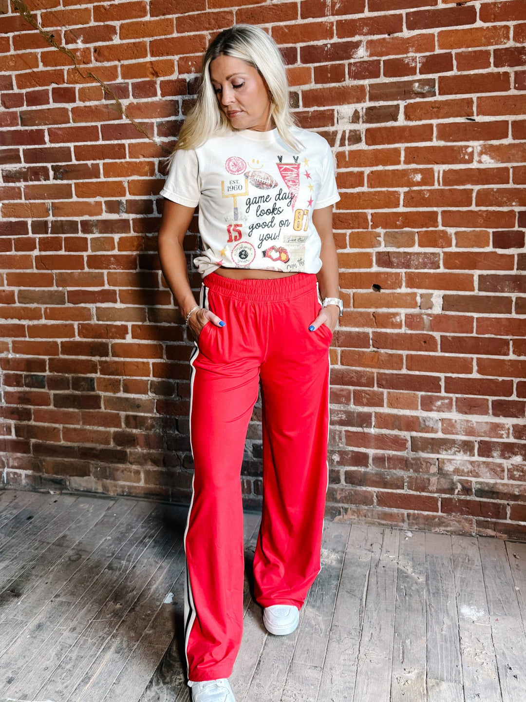 A woman stands against a red brick wall wearing bright red wide-leg sweatpants with white side stripes, a white graphic t-shirt, and white sneakers.