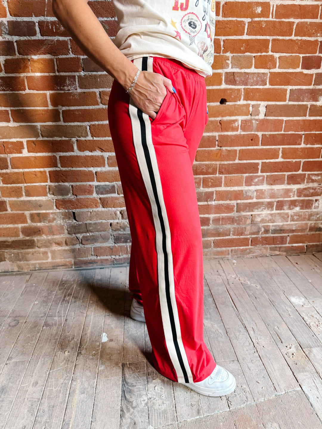 A person wearing bright red wide-leg track pants with a black and white side stripe, standing against a brick wall and wood floor.