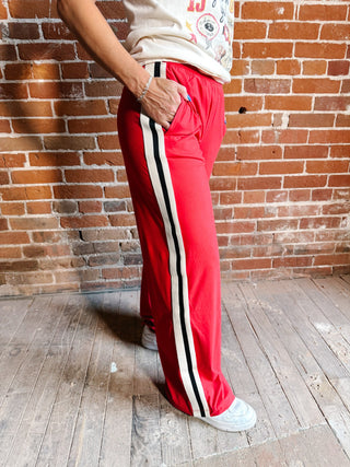 A person wearing bright red wide-leg track pants with a black and white side stripe, standing against a brick wall and wood floor.