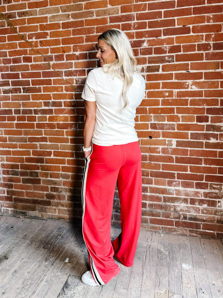 A woman with blonde hair stands with her back to the camera, leaning against a red brick wall. She is wearing bright red wide-leg sweatpants with a white and black side stripe, a white t-shirt, and white sneakers.