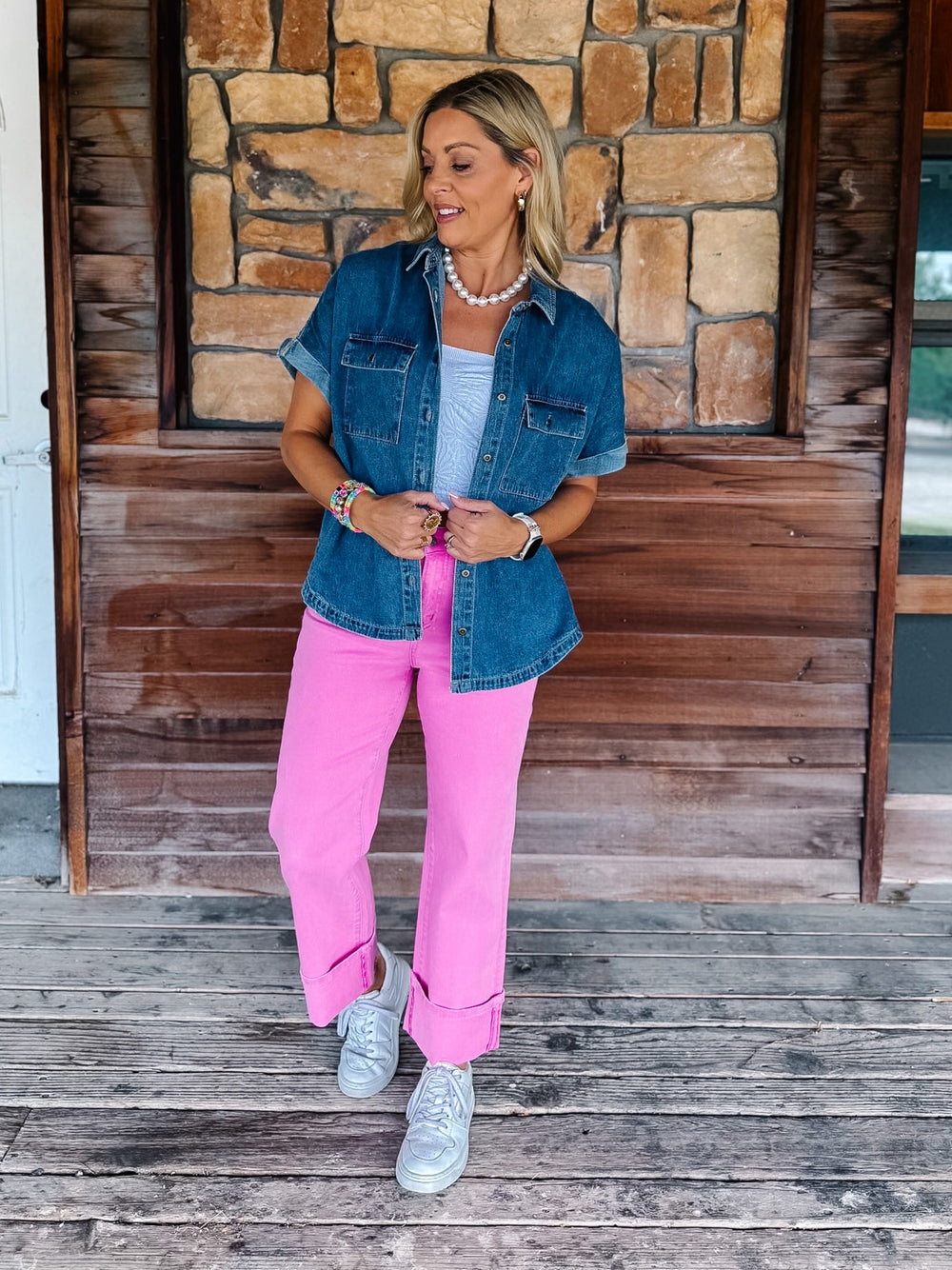 A woman stands on a rustic wooden porch in front of a stone wall, wearing a short-sleeve denim shirt over a white top, bright pink pants, and white sneakers.