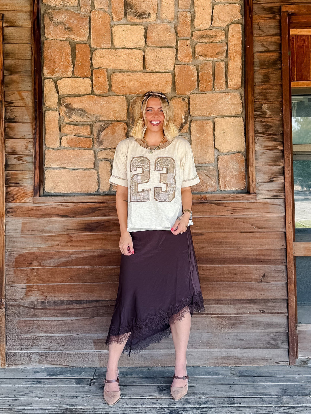 A woman stands on a rustic wooden porch in front of a stone wall, wearing a cream short-sleeve athletic-style top with a large beige &