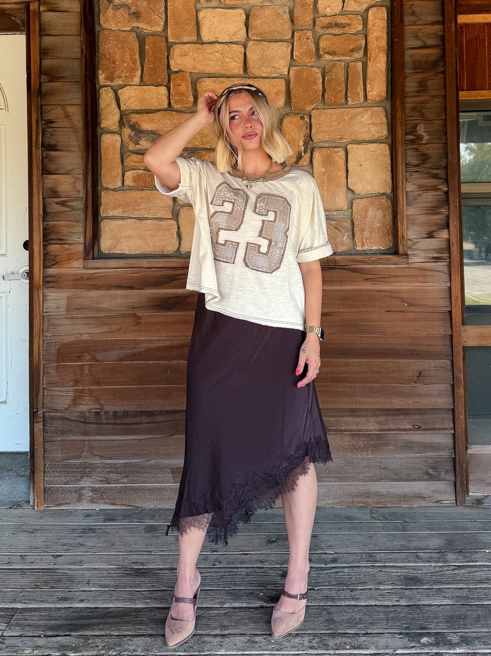 A woman stands on a rustic wooden porch in front of a stone wall, wearing a cream short-sleeve athletic-style top with a large beige '23' graphic and a dark brown midi skirt with a lace-trim hem.