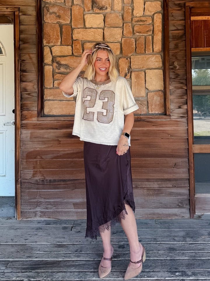 A woman stands on a rustic wooden porch in front of a stone wall, wearing a cream graphic t-shirt with a patchwork number 23 and a dark brown midi skirt.