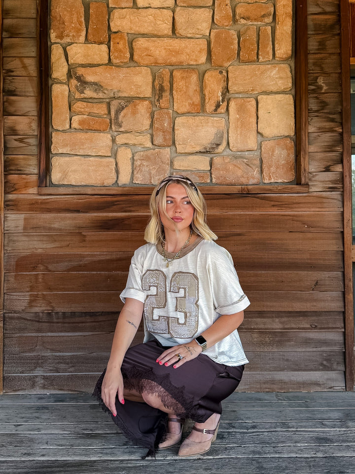 A woman crouching on a rustic wooden porch in front of a stone wall, wearing a white t-shirt with a patchworkn number 23, dark brown skirt, and beige shoes.