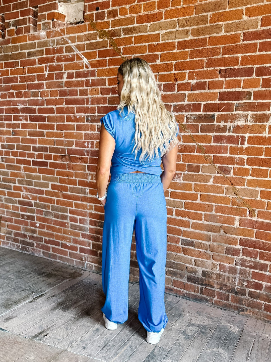 A woman with long blonde hair stands with her back to the camera, wearing a bright blue scrub-style top and matching wide-leg pants, set against a red brick wall and wooden floor.