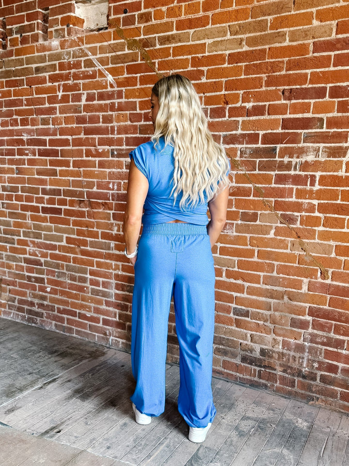 A woman with long blonde hair stands with her back to the camera, wearing a bright blue scrub-style top and matching wide-leg pants, set against a red brick wall and wooden floor.