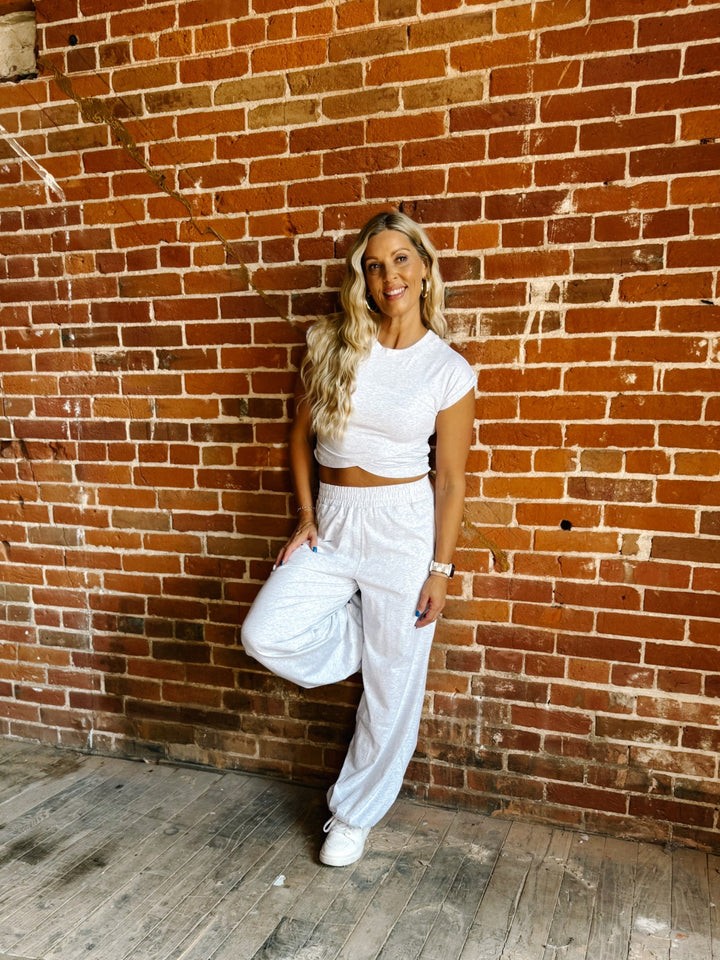 A woman posing in a white two-piece lounge set consisting of a short-sleeve crop top and matching sweatpants, standing against a rustic red-brown brick wall.