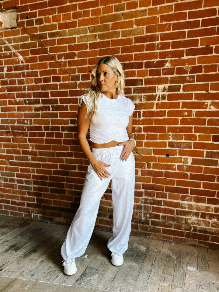 A woman posing in a white two-piece lounge set consisting of a short-sleeve crop top and matching wide-leg sweatpants, set against a rustic red brick wall and wooden floor.