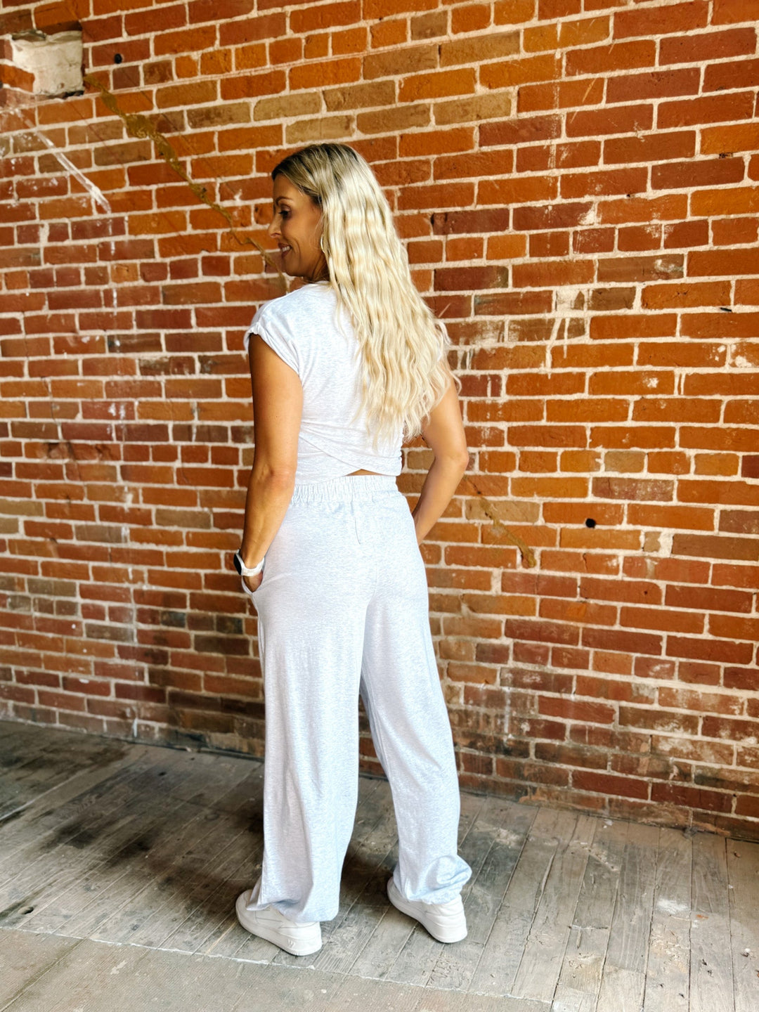 A woman with long blonde hair stands with her back to the camera, wearing a white short-sleeve crop top and matching white wide-leg pants, posing in front of a red brick wall.
