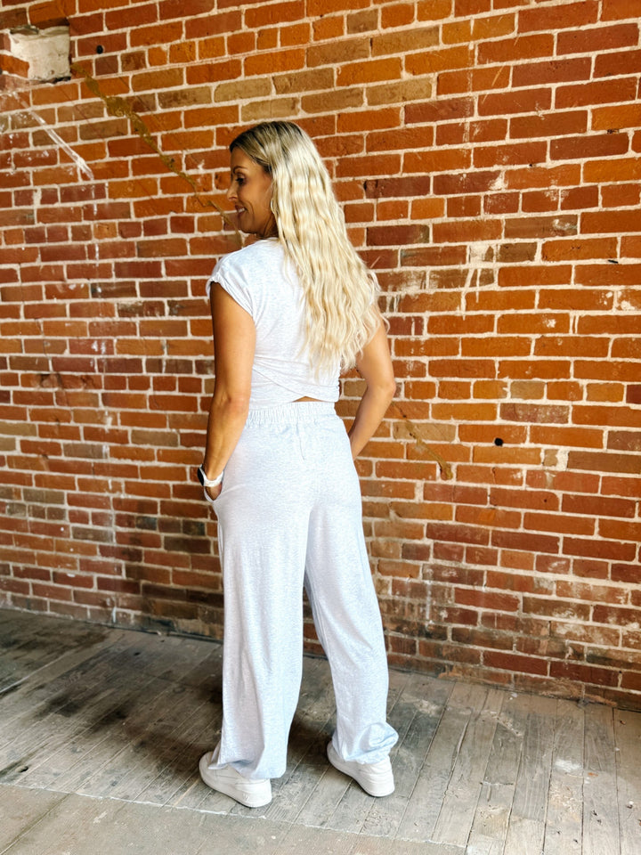 A woman with long blonde hair stands with her back to the camera, wearing a white short-sleeve crop top and matching white wide-leg pants, posing in front of a red brick wall.