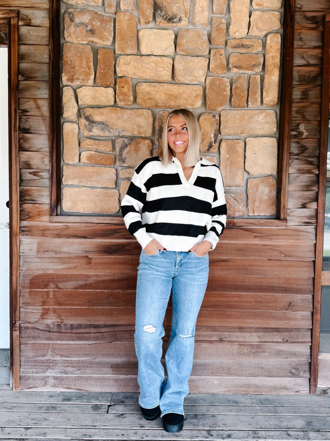 A woman stands on a wooden porch in front of a stone wall, wearing a black and white striped long-sleeve polo sweater, light blue distressed jeans, and black shoes.