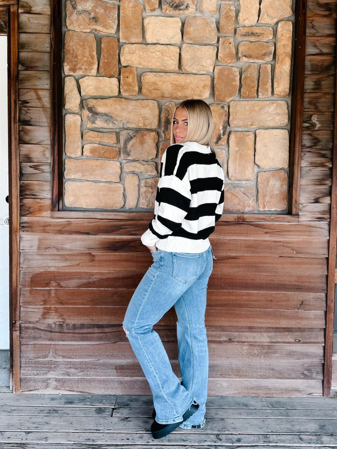 A woman with blonde hair stands on a wooden porch in front of a stone and wood wall, wearing a black and white horizontally striped knit sweater and light blue jeans.
