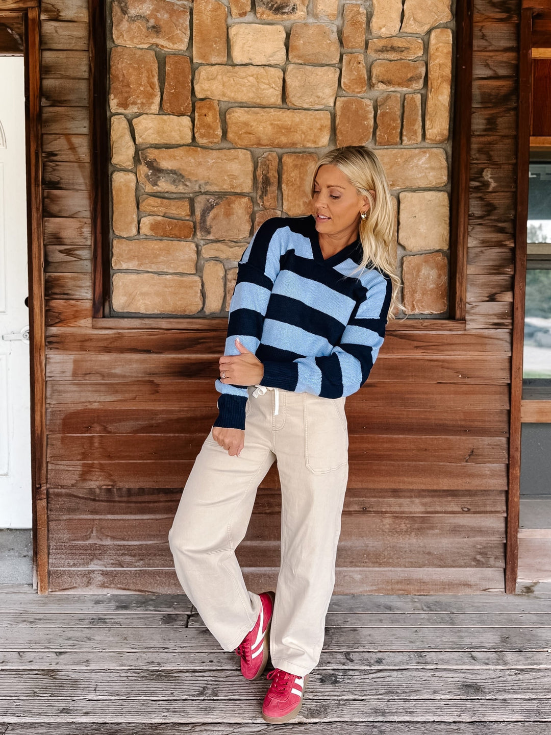 A woman stands on a wooden porch in front of a stone wall, wearing a light blue and navy striped long-sleeve knit sweater, cream wide-leg pants, and bright pink sneakers.