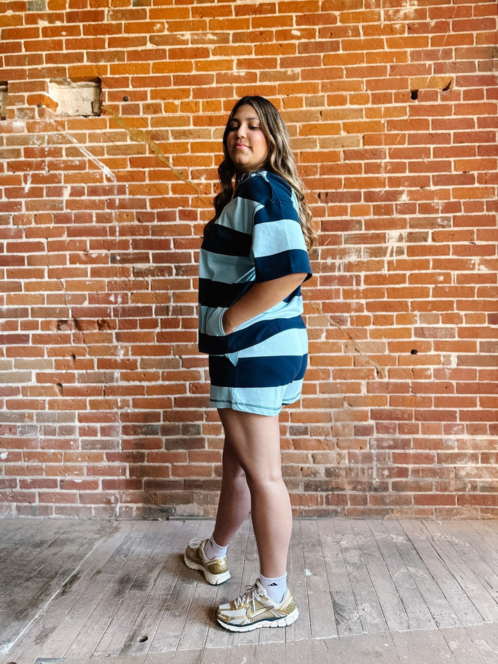 A woman wearing a navy and sky blue striped set with an oversized top and shorts, standing against a brick wall.
