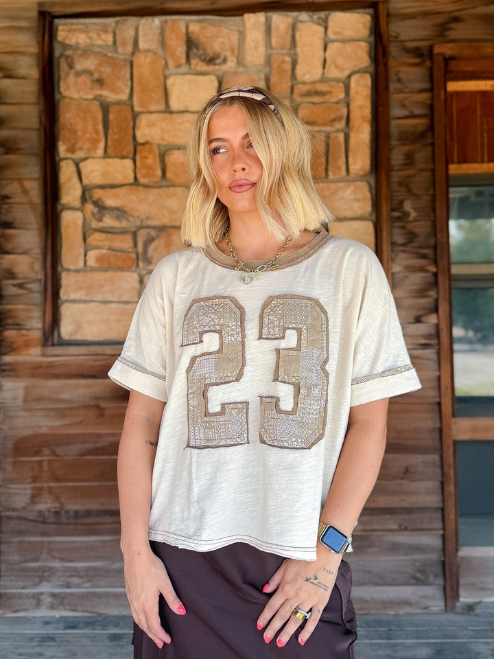 A woman with blonde hair stands in front of a rustic stone and wood wall, wearing a cream-colored short-sleeve t-shirt with a large sequined number 23 on the front, paired with dark brown pants.