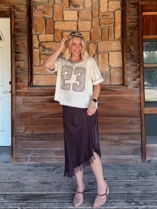 A woman stands on a rustic wooden porch in front of a stone wall, wearing a cream graphic t-shirt with a glittery number 23 and a dark brown midi skirt.