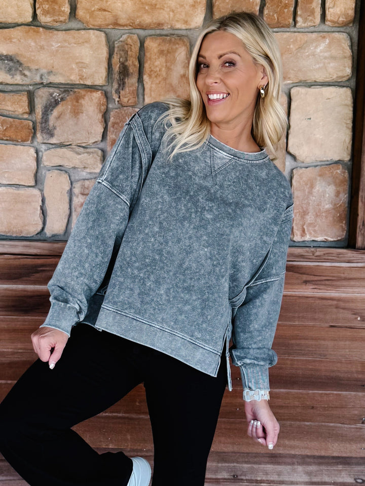 A woman wearing a black acid-wash crewneck sweatshirt and black pants, posing on wooden steps in front of a stone wall.