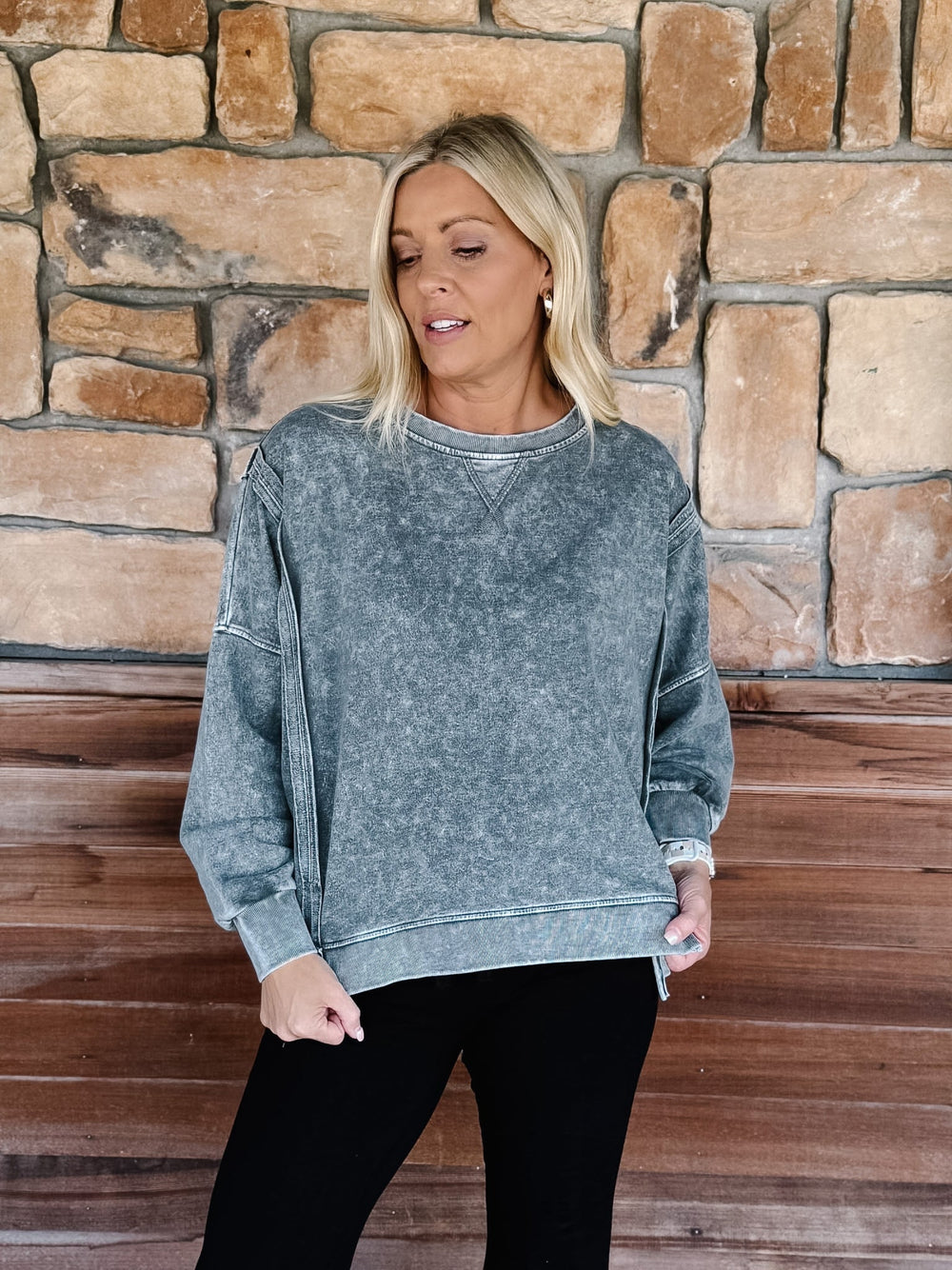 A woman wearing a black acid-wash crewneck sweatshirt and black pants, posing on wooden steps in front of a stone wall.