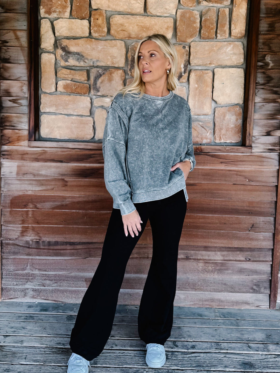 A woman wearing a black acid-wash crewneck sweatshirt and black pants, posing on wooden steps in front of a stone wall.