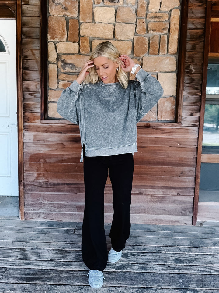 A woman wearing a black acid-wash crewneck sweatshirt and black pants, posing on wooden steps in front of a stone wall.