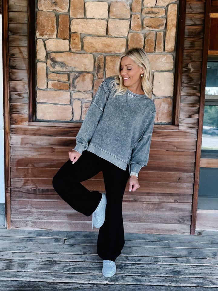 A woman wearing a black acid-wash crewneck sweatshirt and black pants, posing on wooden steps in front of a stone wall.