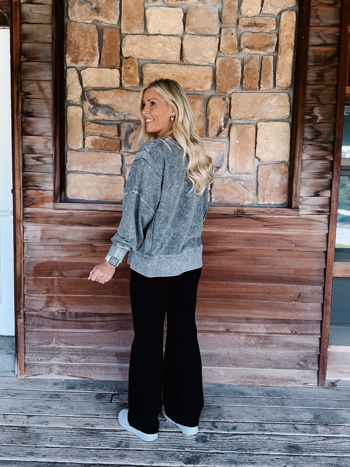 A woman wearing a black acid-wash crewneck sweatshirt and black pants, posing on wooden steps in front of a stone wall.