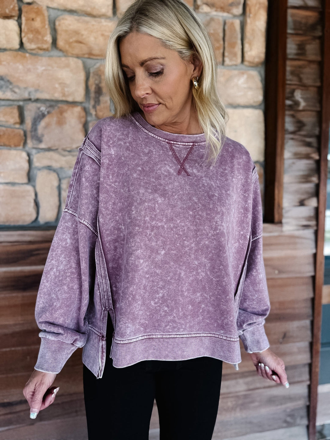 A woman wearing a maroon acid-wash long-sleeve sweatshirt and black pants, standing in front of a rustic stone wall and wooden door.