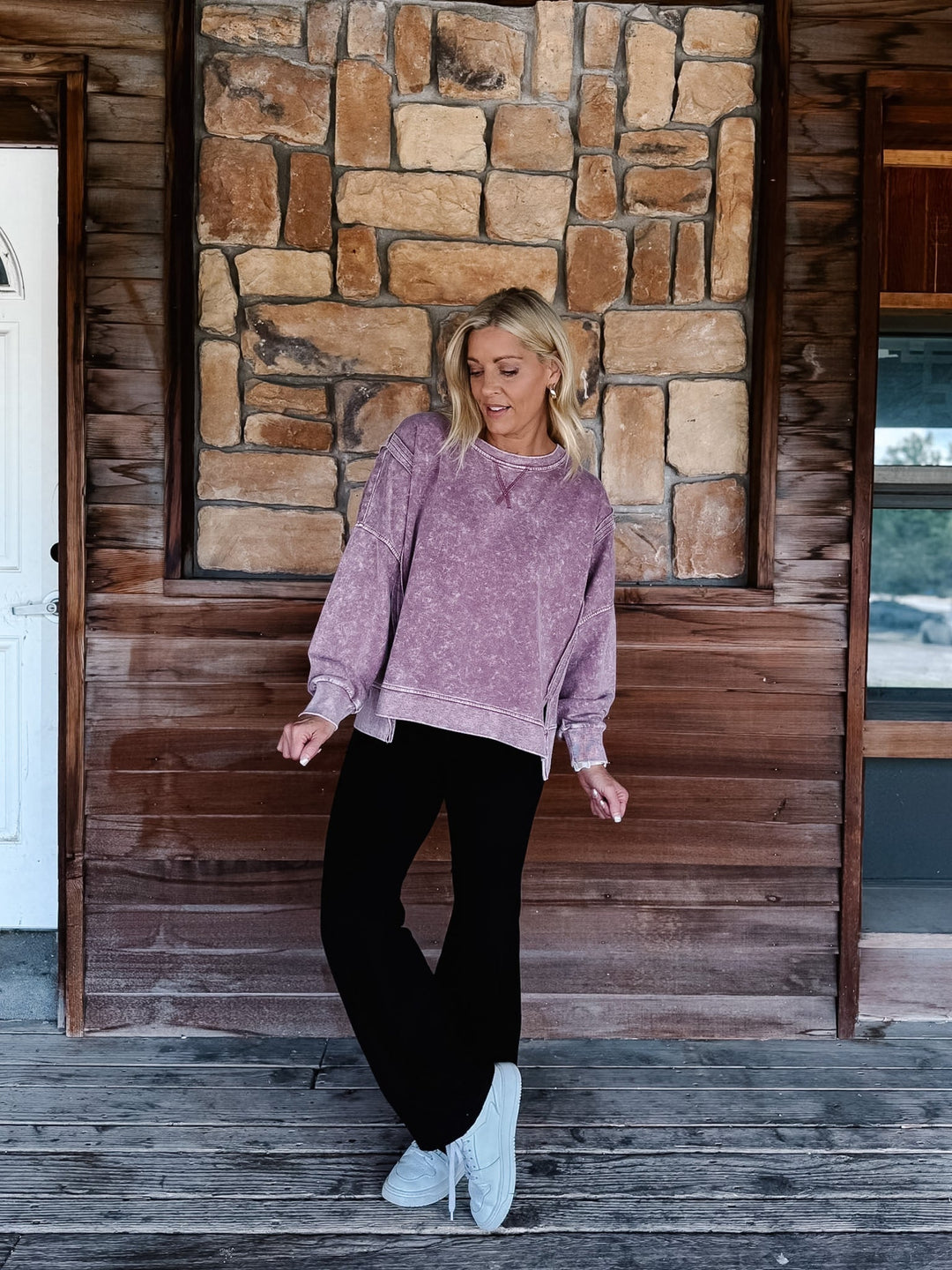 A woman stands on a rustic wooden porch wearing a mineral washed maroon textured crewneck sweatshirt, black pants, and white sneakers.