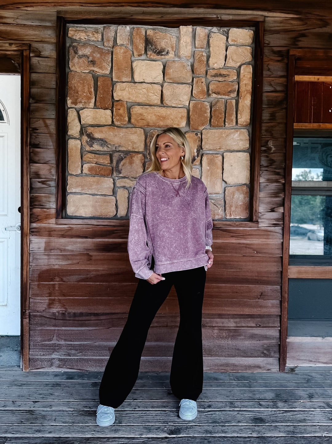 A woman stands on a rustic wooden porch wearing a mineral washed maroon textured crewneck sweatshirt, black pants, and white sneakers.