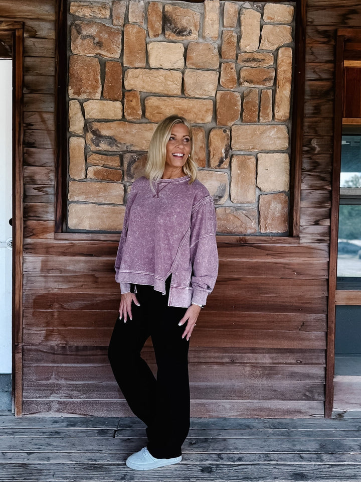 A woman stands on a rustic wooden porch wearing a mineral washed maroon textured crewneck sweatshirt, black pants, and white sneakers.