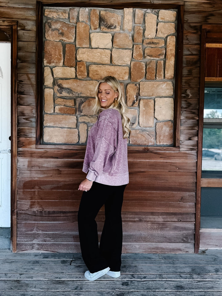 A woman stands on a rustic wooden porch wearing a mineral washed maroon textured crewneck sweatshirt, black pants, and white sneakers.