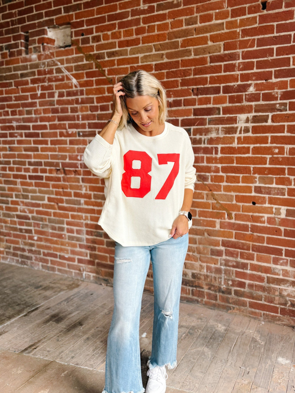 A woman stands in front of a red brick wall wearing a cream-colored long-sleeve sweatshirt with a bold red '87' printed on the front, paired with light-wash distressed jeans and white sneakers.