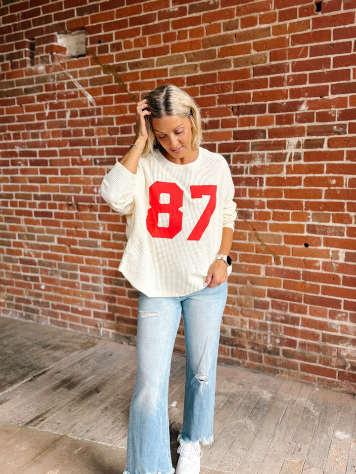 A woman stands in front of a red brick wall wearing a cream-colored long-sleeve sweatshirt with a bold red &