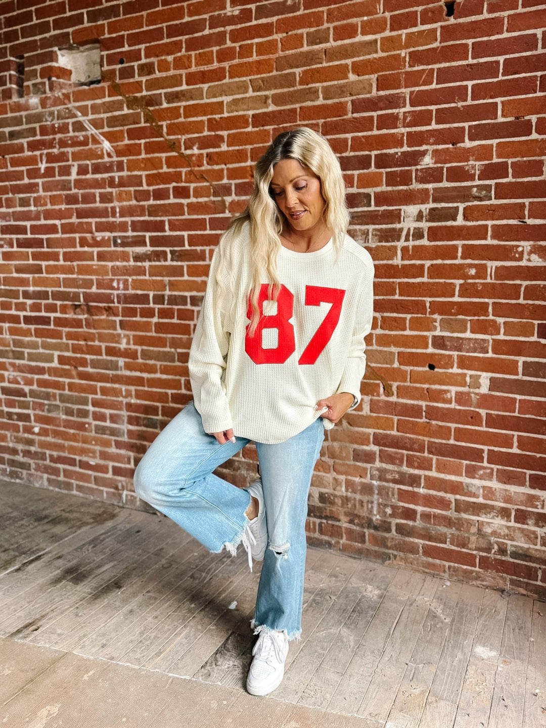 A woman posing in front of a brick wall wearing a cream-colored crewneck sweatshirt with a bold red &