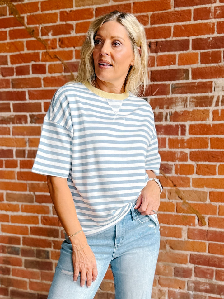 A woman wearing a light blue and white striped short-sleeve t-shirt with a yellow crew neck trim, paired with light-wash blue jeans, standing in front of a red brick wall.
