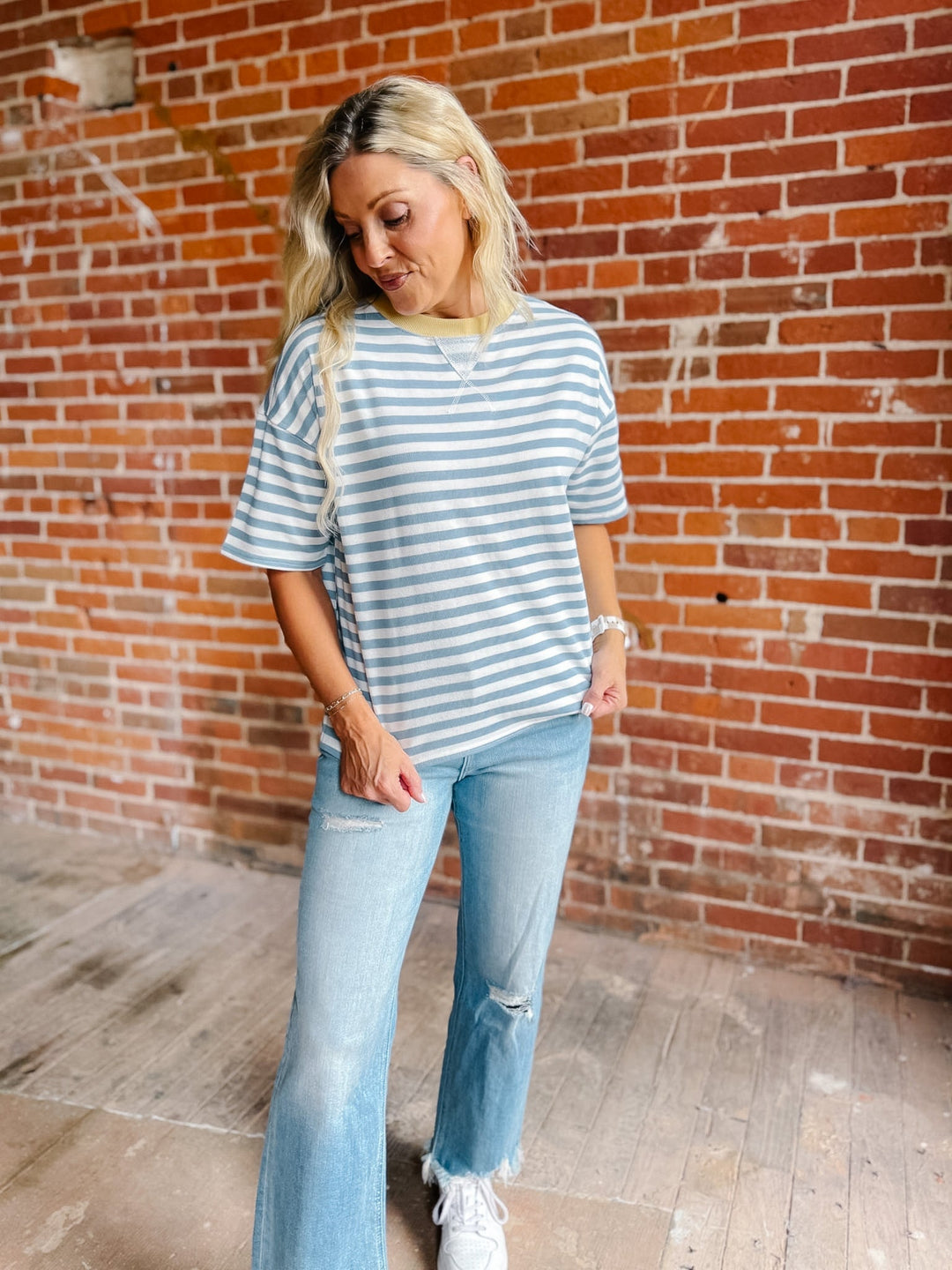 A woman wearing a light blue and white striped short-sleeve t-shirt, light-wash blue jeans, and white sneakers, standing in front of a red brick wall.