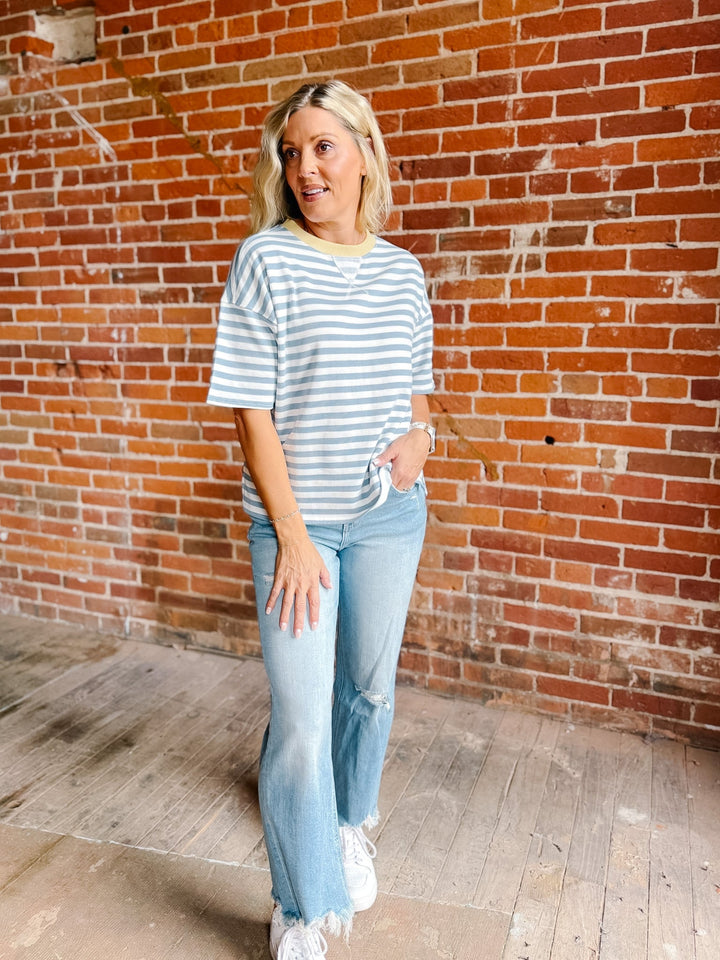 A woman wearing a light blue and white striped t-shirt, light-wash distressed jeans, and white sneakers, standing in front of a red brick wall.