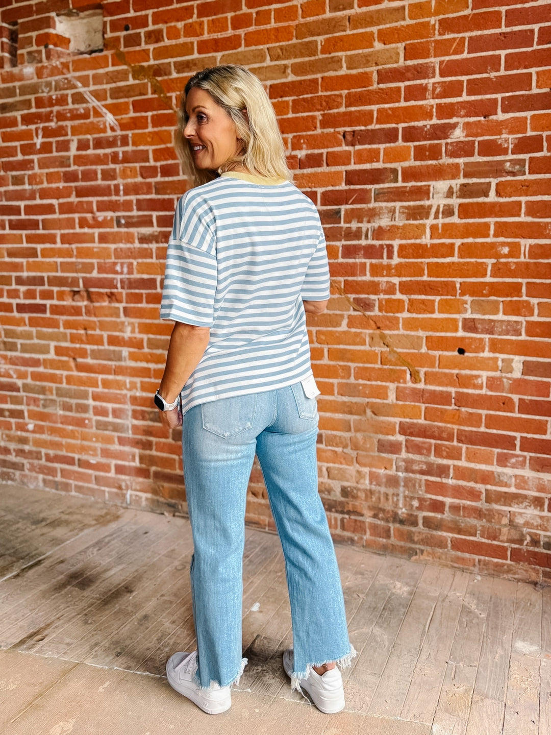 A woman with blonde hair stands in front of a red brick wall, wearing a light blue and white striped t-shirt, light-wash blue jeans, and white sneakers.