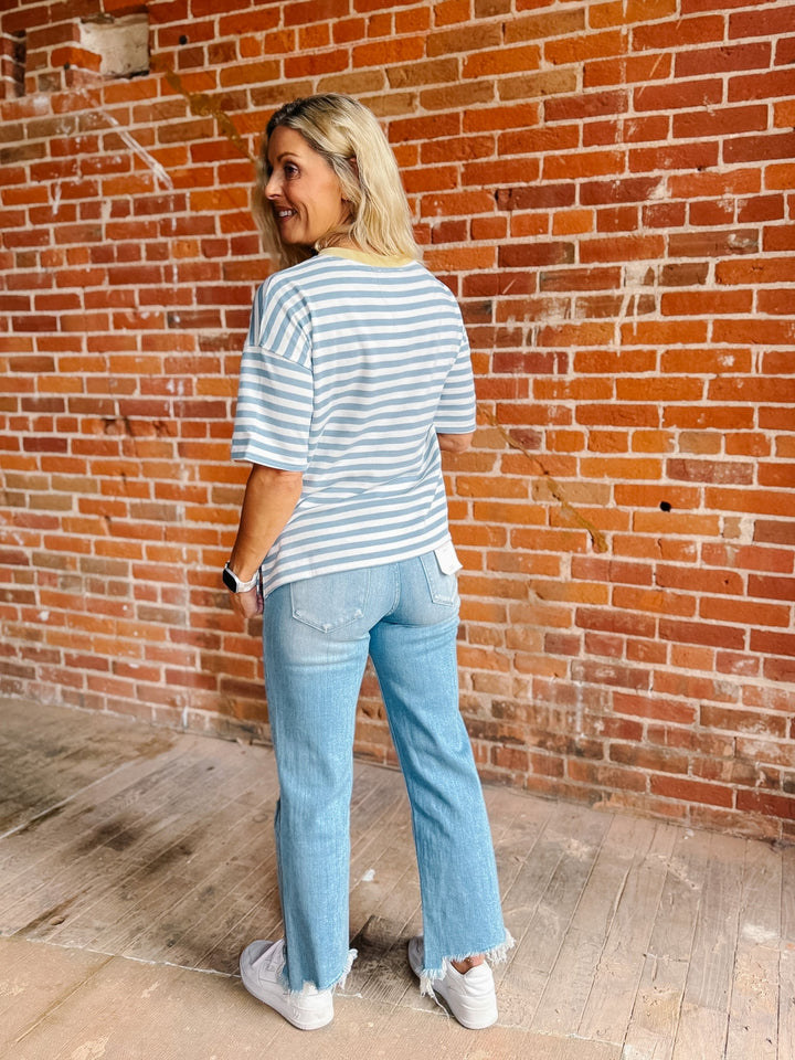 A woman with blonde hair stands in front of a red brick wall, wearing a light blue and white striped t-shirt, light-wash blue jeans, and white sneakers.